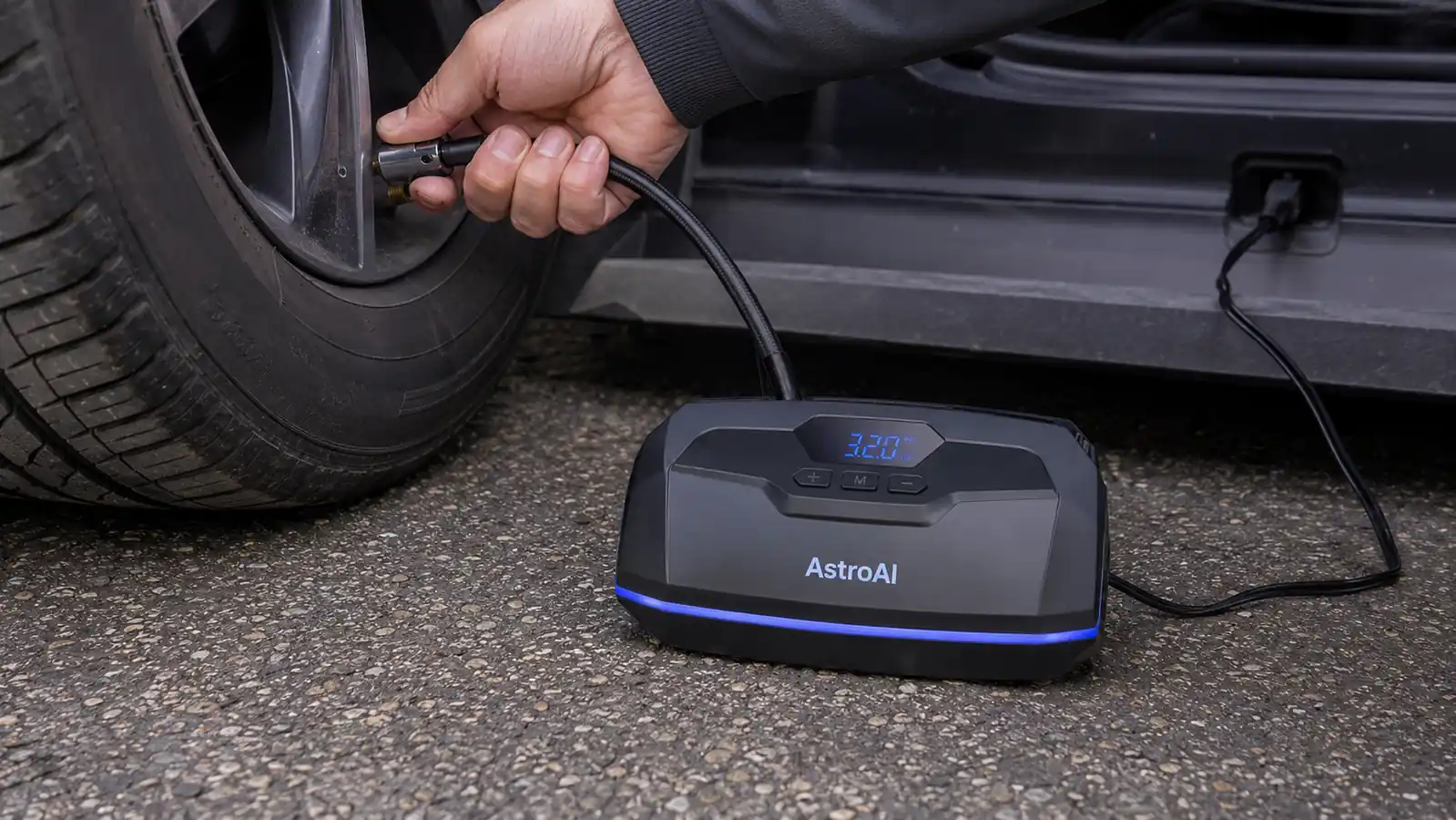 Hand holding the AstroAI tire inflator nozzle against a car tire valve stem, digital display lit up