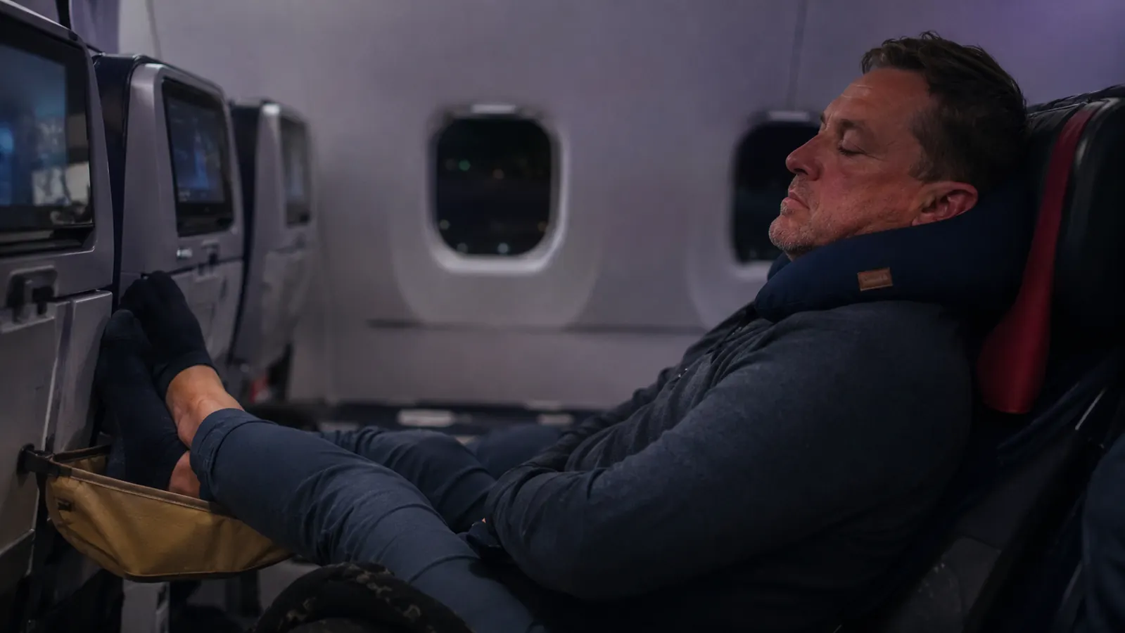 Traveler asleep on a long flight with feet elevated on a footrest hammock and a travel pillow behind their head