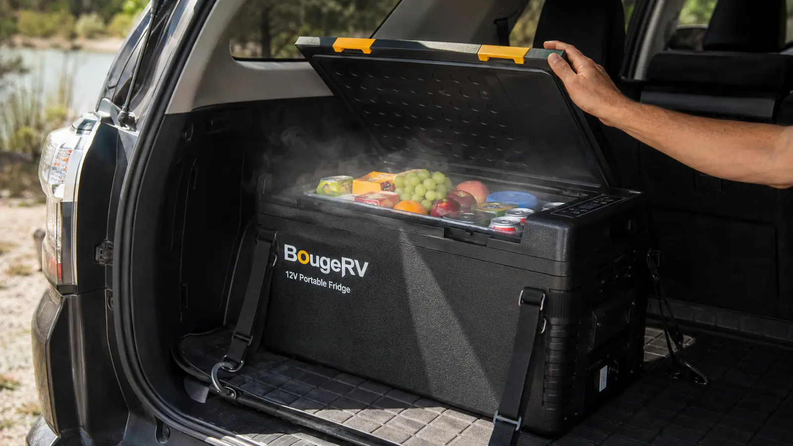 Hand lifting the lid of a black portable 12V fridge loaded in the cargo area of an SUV, revealing cold drinks and packed food inside