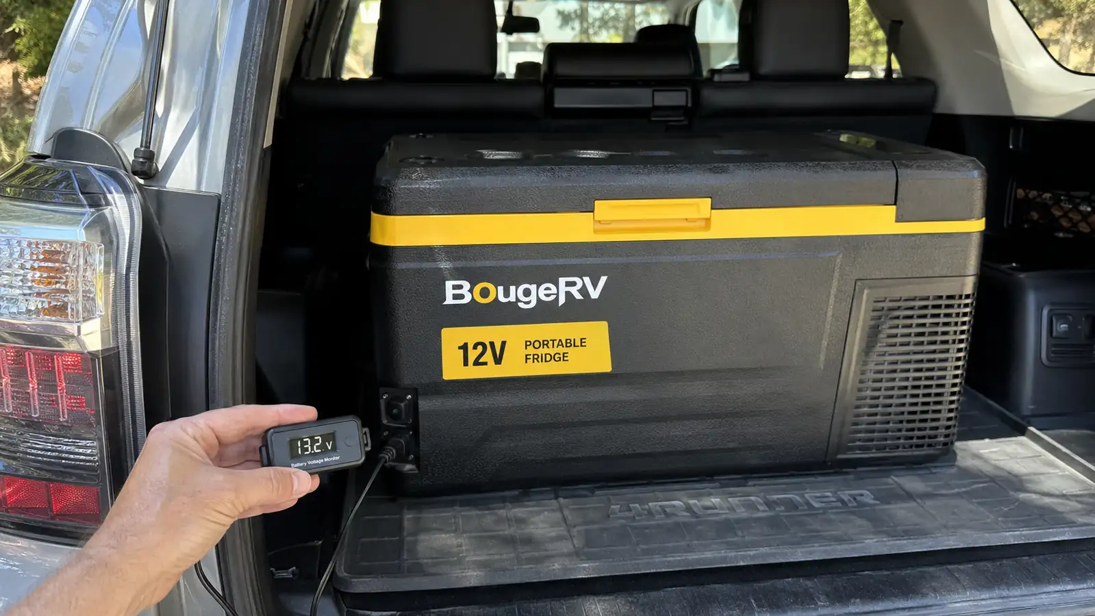 Hand holding a small battery monitor next to the BougeRV fridge's power cable in the back of a 4Runner