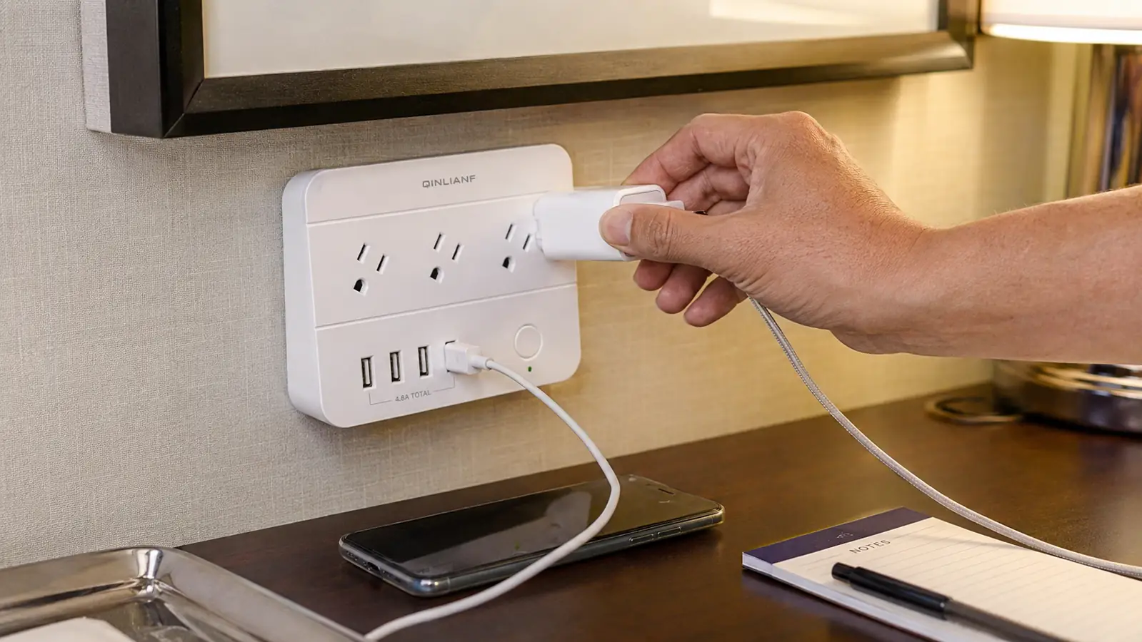 Hand plugging a laptop charger into the QINLIANF power strip on a hotel desk with a phone already charging on a USB port