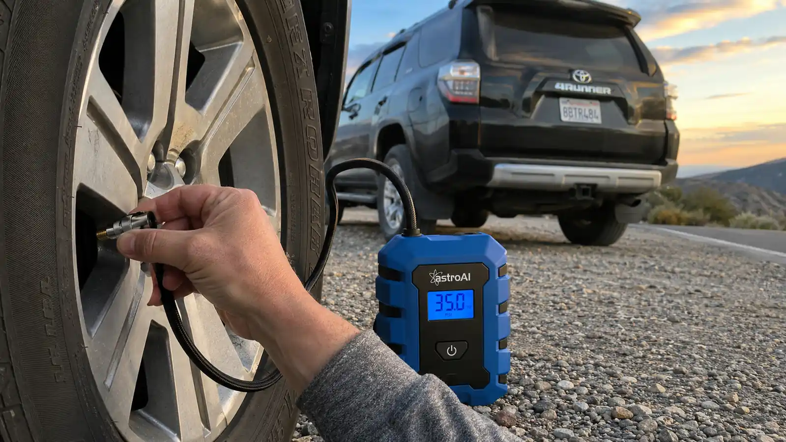 Hand holding a blue portable tire inflator with digital gauge up to a car tire's valve stem on the shoulder of a rural highway